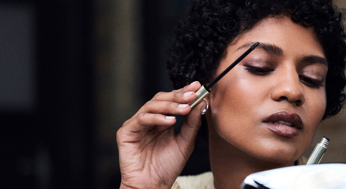 A close-up lifestyle shot of a woman with curly black hair applying eyebrow pomade with a spoolie wand, looking into a mirror.