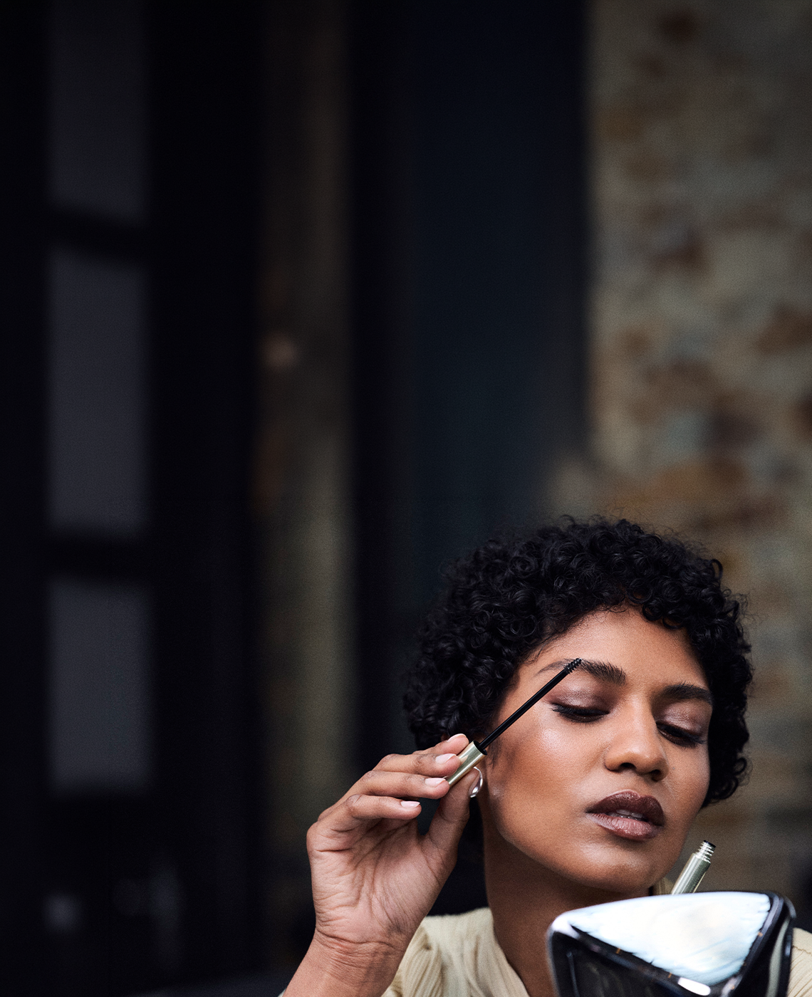A close-up lifestyle shot of a woman with short curly black hair applying dark brown eyebrow pomade with a spoolie, holding a small mirror in her other hand.